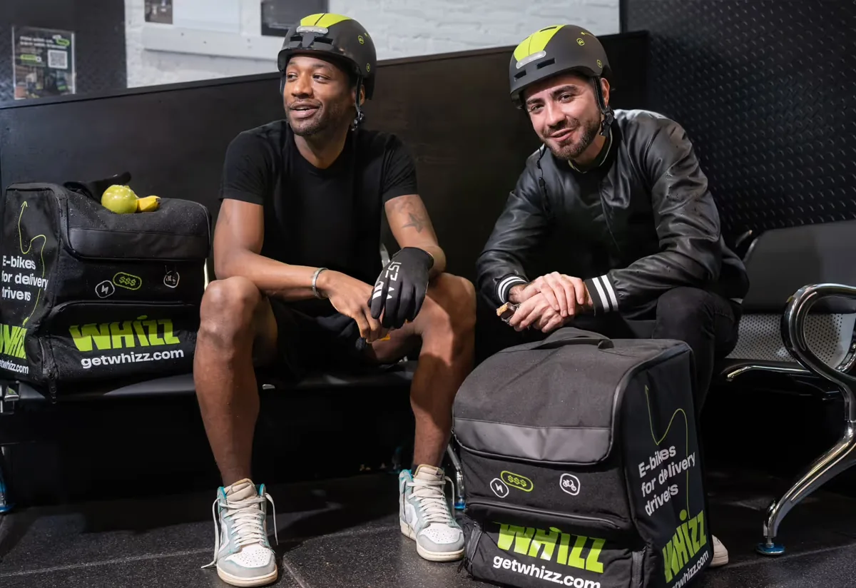 Two Whizz riders in helmets laughing with their delivery bags