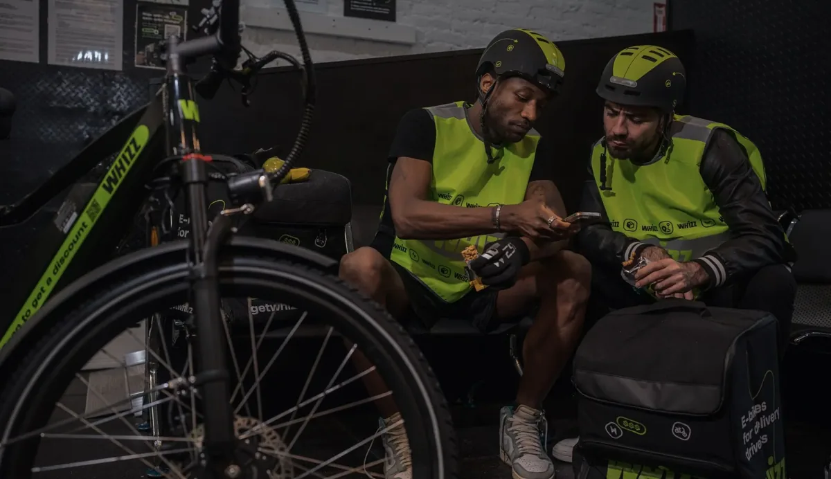 Two Whizz riders in safety vests checking a delivery bag before a shift